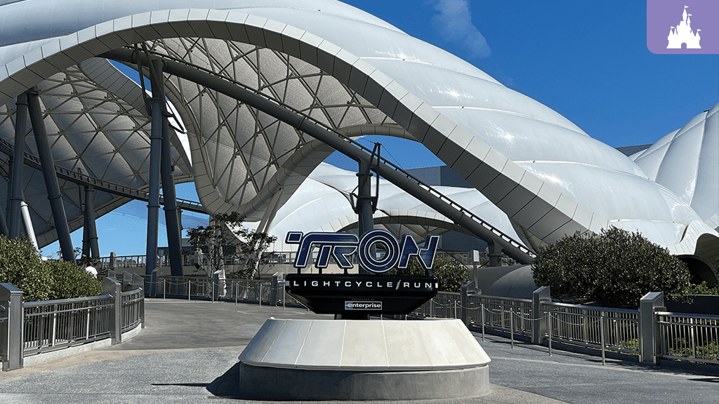 A futuristic entrance with the "TRON Lightcycle Run" sign underneath a sleek, white, curved structure against a blue sky.