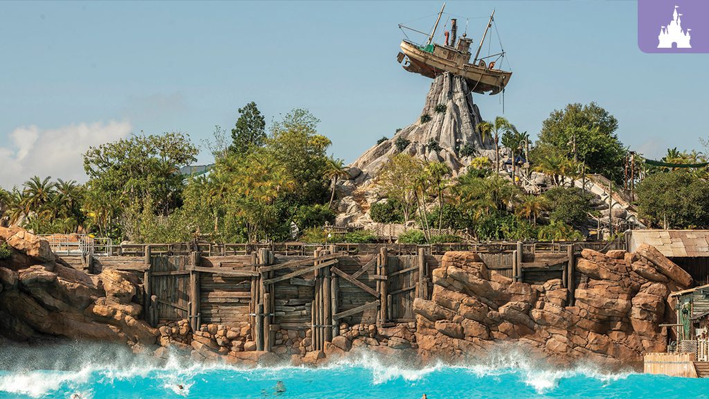 A scenic view of a water park featuring a rocky mountain with a ship on top, surrounded by lush greenery and a bright blue pool.