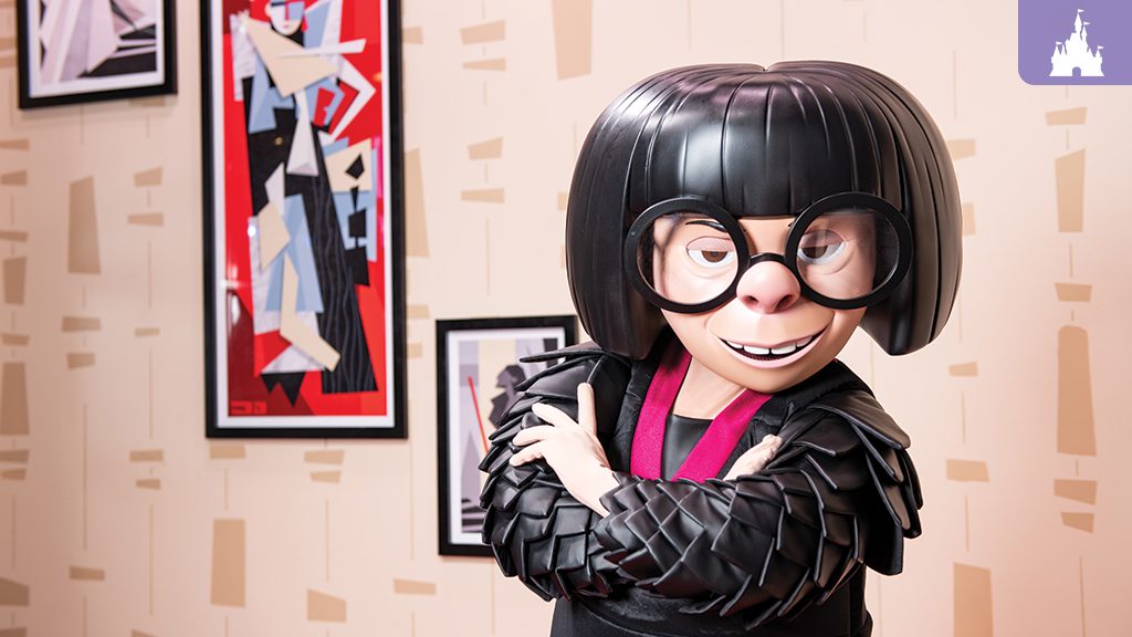 A character with black bobbed hair and large glasses stands confidently, wearing a stylish black outfit against a colorful wall of art.