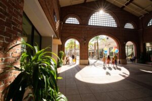 A bright, airy indoor space with an arched brick design, palm trees, and people walking, illuminated by sunlight streaming through windows.