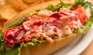 A lobster roll filled with creamy lobster meat and lettuce, served in a toasted bun, accompanied by potato chips in the background.