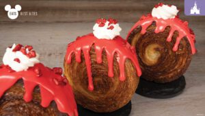 Three swirled pastries topped with red icing and whipped cream, resembling festive desserts, displayed on a wooden surface.