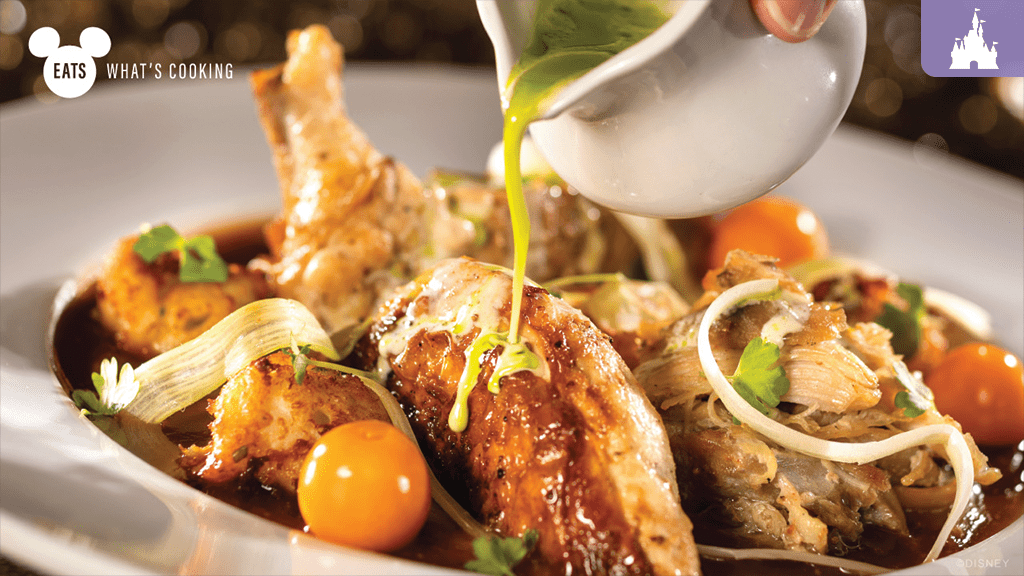 A close-up of a plated dish featuring pieces of chicken, cherry tomatoes, and herbs, with sauce being drizzled over the top.