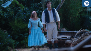 A girl in a blue dress and a young man in a vest stand together in a lush, green outdoor setting, looking ahead.