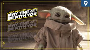 A cute, large-eared character in a beige robe stands against a backdrop featuring "Star Wars Day: May the 4th Be With You" text.