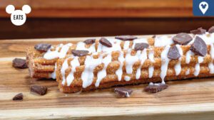 Two churros drizzled with white icing and sprinkled with chocolate chips, displayed on a wooden platter.