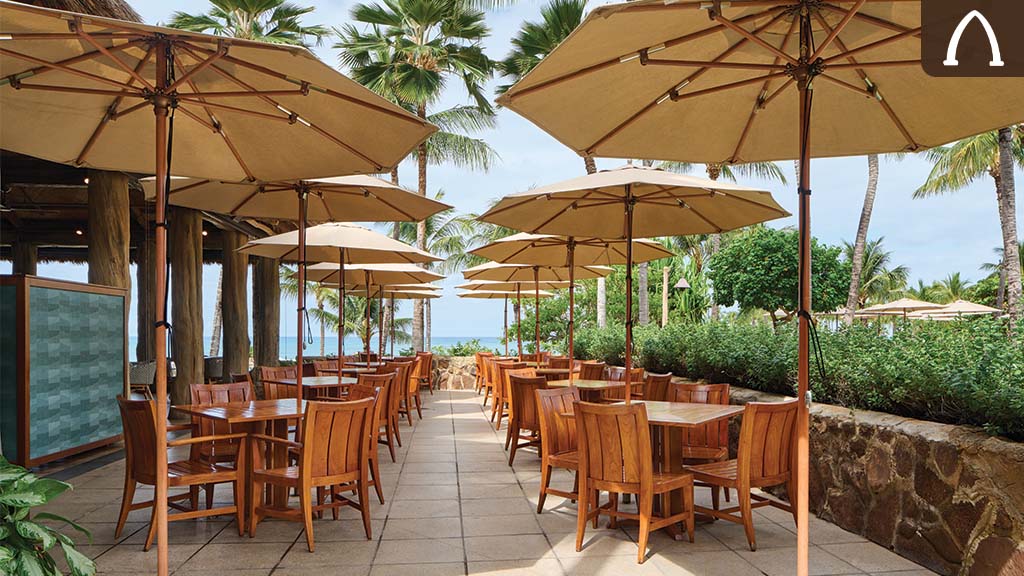 Outdoor dining area with wooden tables and umbrellas, surrounded by palm trees and greenery, overlooking a tranquil beach.