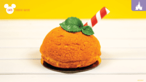 A vibrant orange dessert shaped like a sphere, topped with a mint leaf and a red-and-white striped straw, against a bright yellow background.