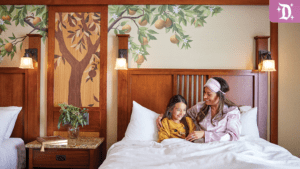 A mother and daughter cuddle in bed, surrounded by warm decor and a mural of a tree, creating a cozy, inviting atmosphere.