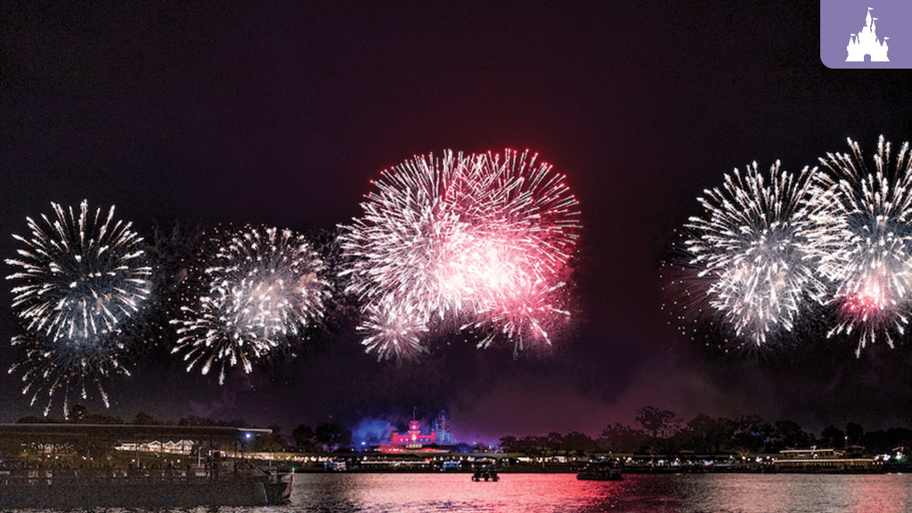 magic-kingdom-july-4-military-flyover Colorful fireworks illuminate the night sky over a serene body of water, with a distant castle silhouette in the background.