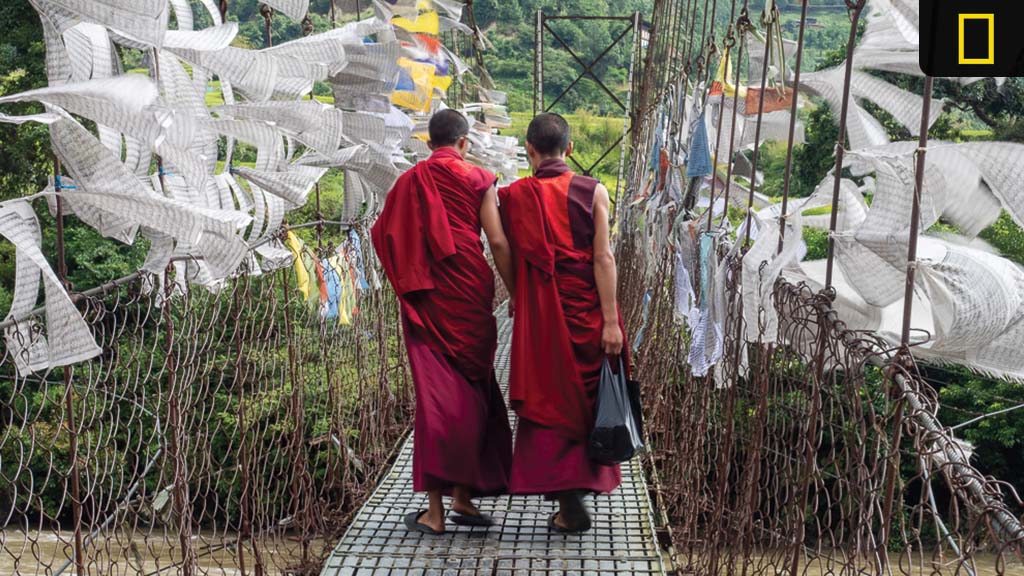 nat-geo-new-countries-and-itineraries Two monks in red robes walk across a swinging bridge adorned with prayer flags, surrounded by lush greenery.