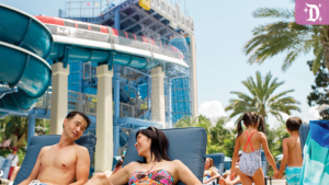 A couple relaxes in lounge chairs by a water park, with a large water slide and palm trees in the background. Kids play nearby.