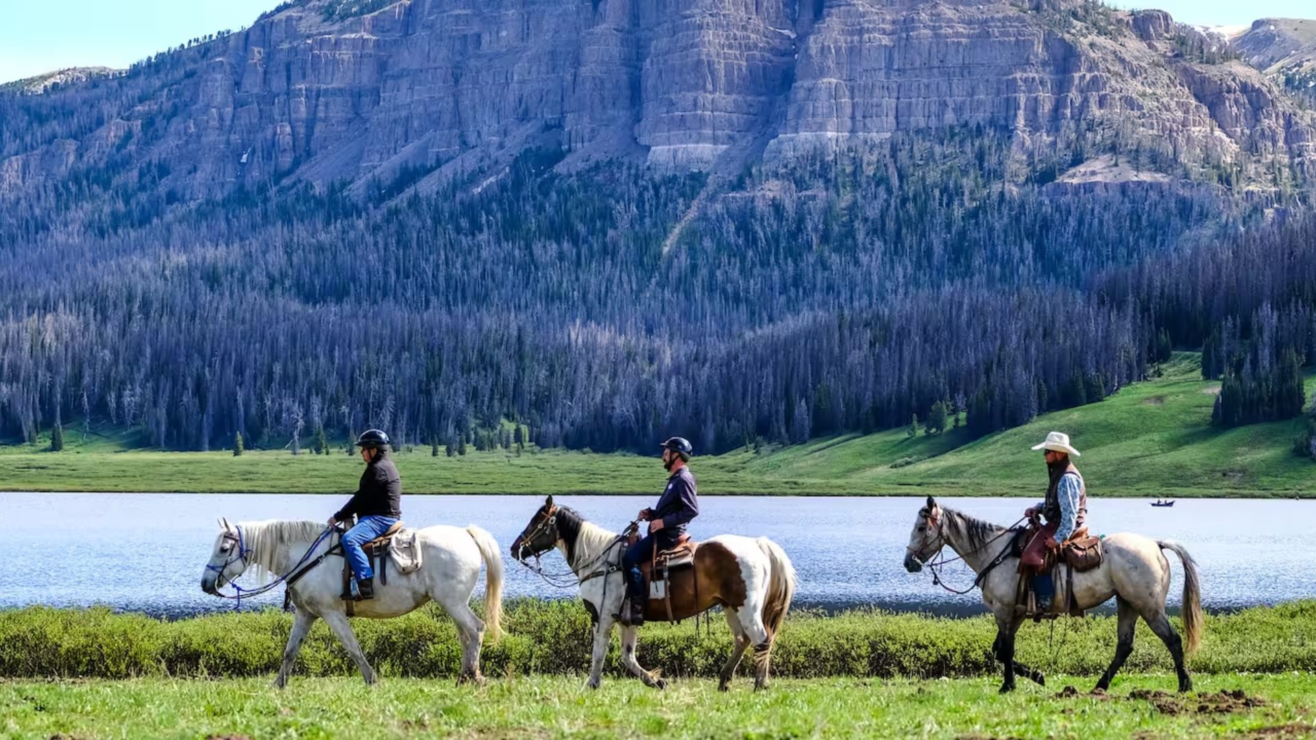 AdventuresByDisneyWyoming Three riders on horseback traverse a lush green field beside a tranquil lake, with a majestic mountain backdrop.