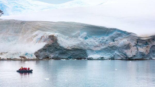 AdventuresbyDisneyAntarcticaandPatagoniaExpeditionCruise A small group of people in red jackets travels on a raft near a glacier in calm, icy waters. The landscape features icy blue and white hues.