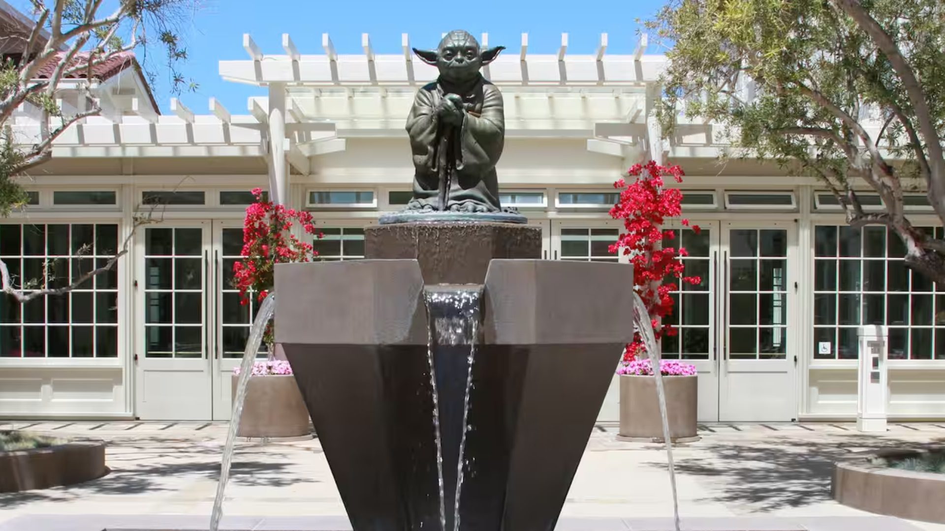 AdventuresbyDisneyAroundtheWorldPrivateJetExperienceLucasFilmCampus A bronze statue of a small character stands in a water fountain, surrounded by red flowers and a building in the background.