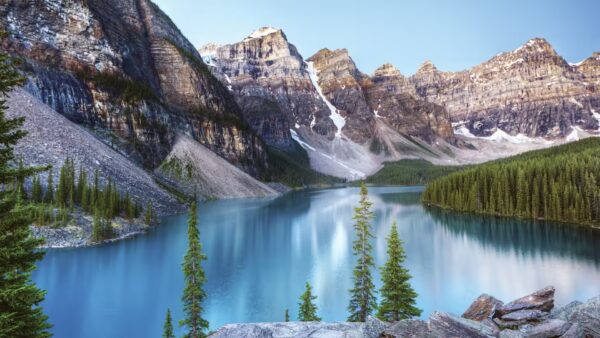 AdventuresbyDisneyCanadianRockies A serene mountain landscape featuring a calm lake surrounded by dense evergreen forests and towering rocky peaks under a clear blue sky.