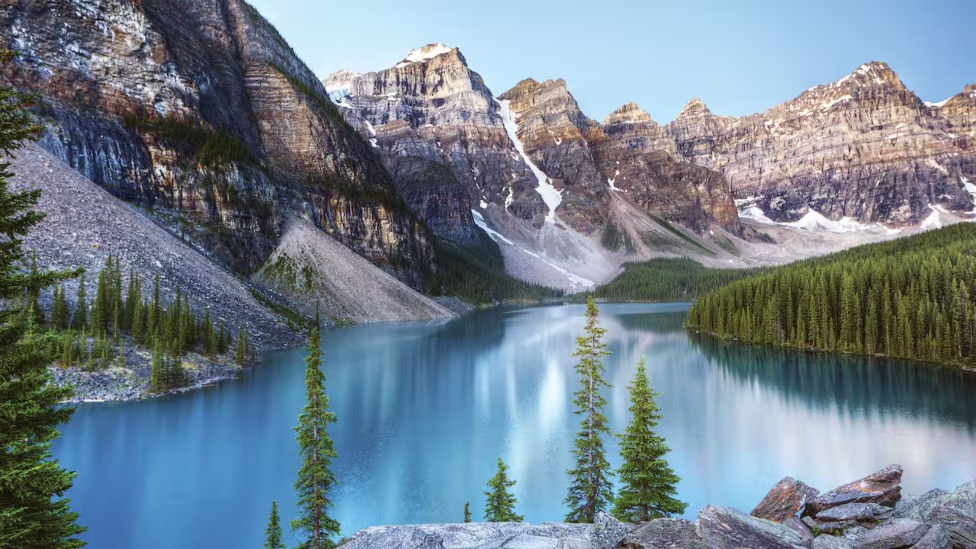 A serene mountain landscape featuring a calm lake surrounded by dense evergreen forests and towering rocky peaks under a clear blue sky.