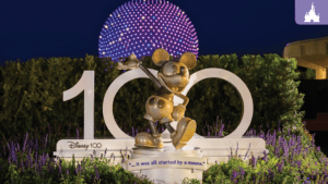A bronze statue of Mickey Mouse stands in front of a large "100" sign, illuminated at night with colorful lights in the background.