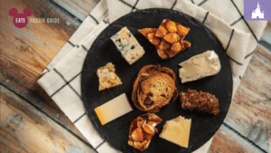 A black slate platter features an assortment of cheeses, crunchy snacks, and a pastry, arranged neatly on a light checkered cloth.