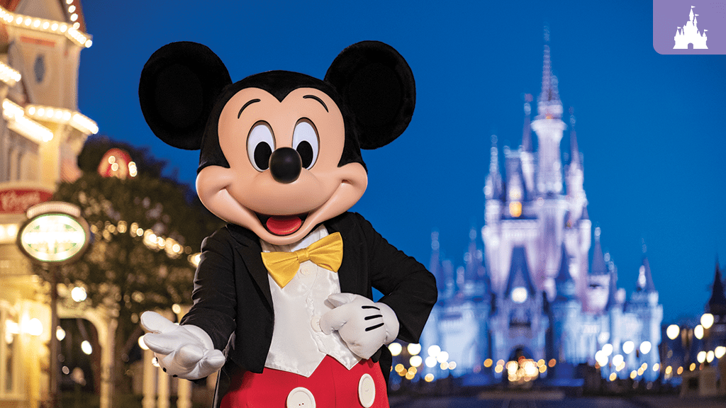 Mickey Mouse, dressed in a black tuxedo with a yellow bow tie, poses cheerfully in front of a brightly lit Cinderella Castle at night in a Disney theme park.