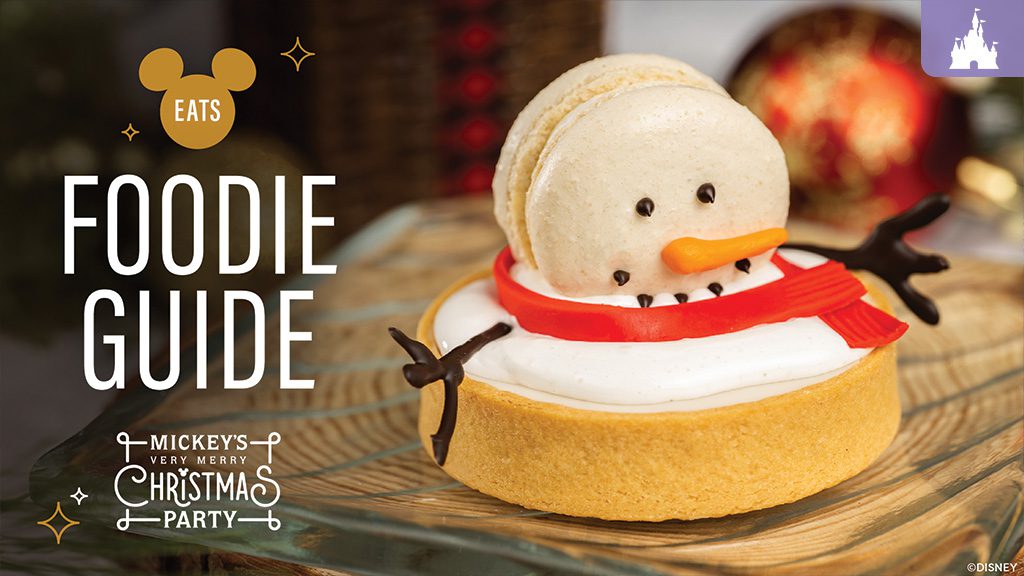 2025-mickeys-very-merry-christmas-party-foodie-guide A festive tart decorated like a snowman, featuring a macaron for the head, black icing eyes, a carrot-shaped nose, chocolate stick arms, and a red candy scarf, sitting on a wooden surface. Text reads "Foodie Guide" for Mickey's Very Merry Christmas Party.