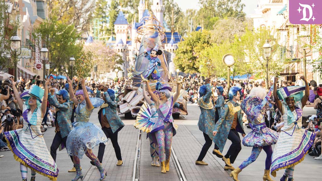 Colorful dancers in patterned outfits lead a street parade past cheering crowds, flanked by a float with a cartoon mouse figure and a castle in view