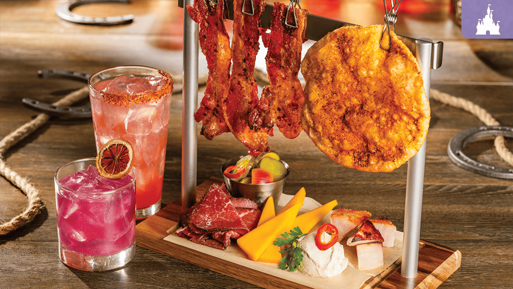 Seasoned bacon strips and a round flatbread hang over a board with cheese wedges, sliced cured meats, pickles, chili dip, and two pink cocktails
