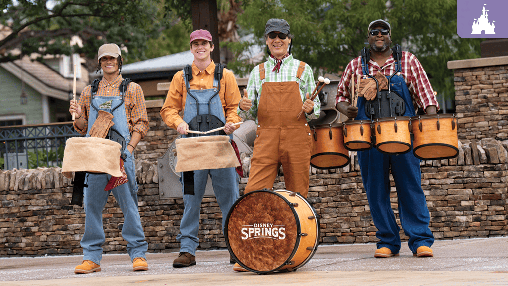 Four drummers in caps and overalls stand outdoors by a stone wall, each holding mallets and wooden drums, including a bass drum labeled Disney Springs