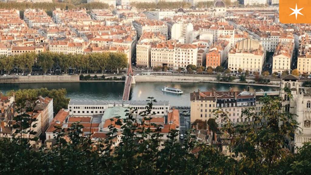 A scenic view of Lyon, France, showcasing the Saône River, a pedestrian bridge, and historic buildings with red-tiled roofs. A river cruise boat is sailing through the water.