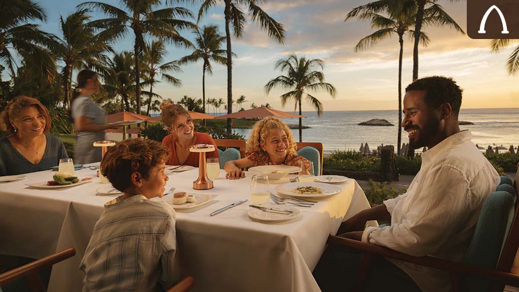 Family dining outdoors at Aulani Resort at sunset with ocean views, palm trees, and a tropical setting in the background.