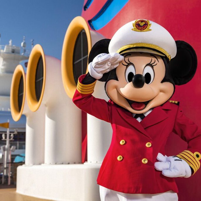 Minnie Mouse dressed in a captain’s uniform salutes, standing on a cruise ship deck with colorful structures in the background.