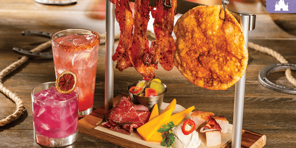 Seasoned bacon strips and a round flatbread hang over a board with cheese wedges, sliced cured meats, pickles, chili dip, and two pink cocktails