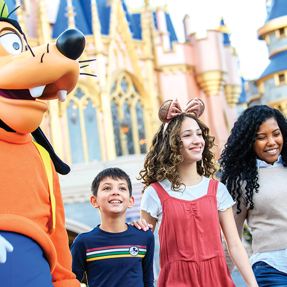 A cheerful Goofy walks alongside a smiling family, including a boy and three adults, in front of a colorful castle at a theme park.