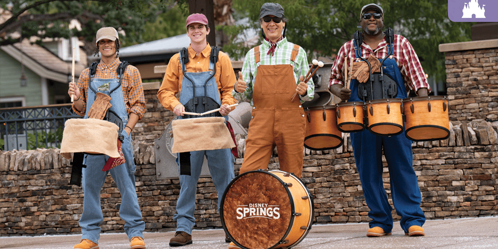 Four drummers in caps and overalls stand outdoors by a stone wall, each holding mallets and wooden drums, including a bass drum labeled Disney Springs