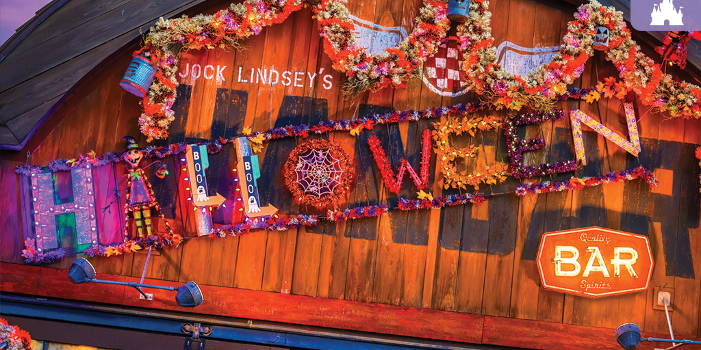 Wooden bar facade reading Jock Lindsey’s HALLOWEEN BAR, decorated with autumn garlands, a small scarecrow figure and a glowing neon BAR sign