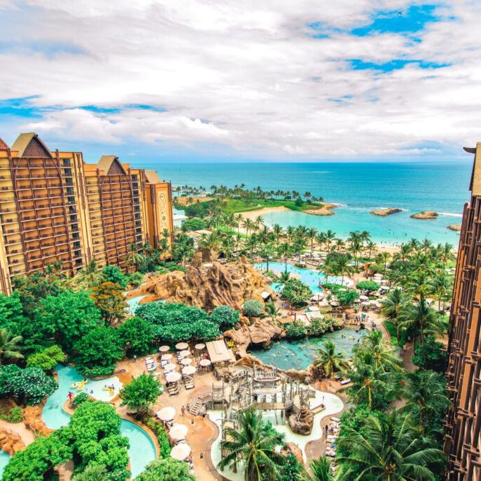 Aerial view of a tropical resort with green landscaping, pools, and the ocean in the background under a partly cloudy sky.