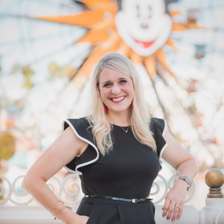 Brittney-Poe A smiling woman in a black dress stands with hands on hips, with a colorful Mickey Mouse Ferris wheel in the background.