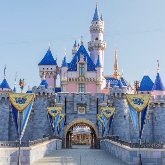 A fairy-tale castle with blue turrets and pink walls, adorned with banners, set against a clear sky. Pathway leads to the entrance.