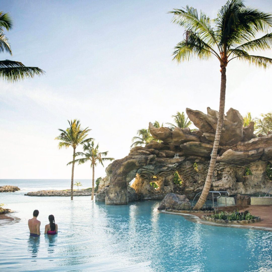 Two people stand in a tranquil pool surrounded by palm trees, with a rocky formation and ocean in the background, under a sunny sky.