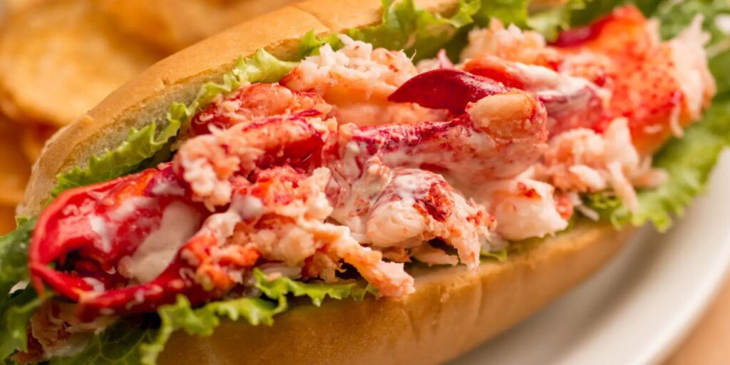 A lobster roll filled with creamy lobster meat and lettuce, served in a toasted bun, accompanied by potato chips in the background.