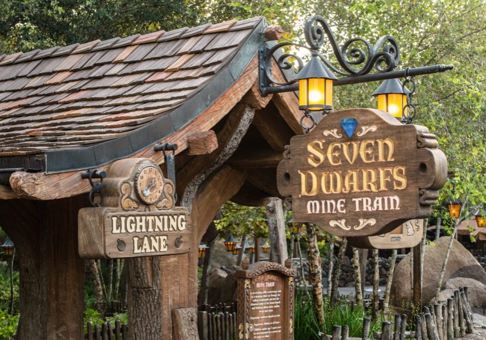 MagicKingdomSevenDwarfsMineTrain Sign for "Seven Dwarfs Mine Train" attraction with an arrow for "Lightning Lane," surrounded by greenery and rustic structures.