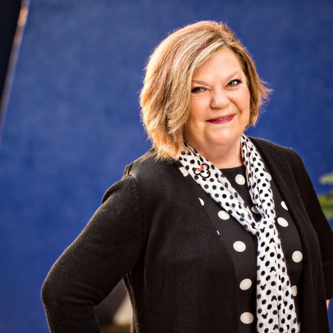 A smiling woman stands confidently in front of a blue backdrop, wearing a black cardigan and a polka dot scarf. Green foliage is visible nearby.