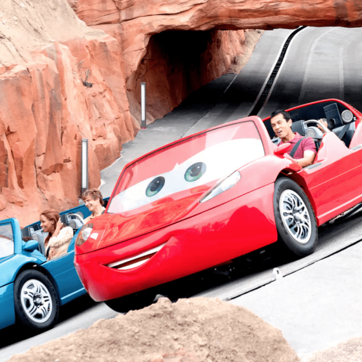 Two animated cars, one red and one blue with large eyes, are riding on a winding track through a rocky landscape.