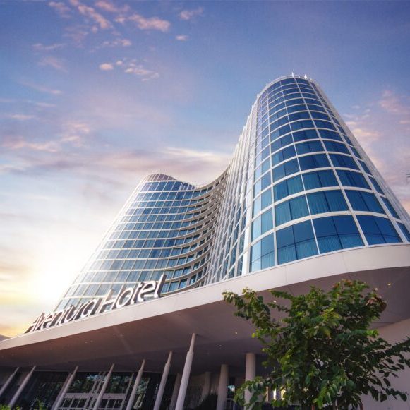 Modern hotel with a sleek glass facade, set against a vibrant sky, framed by palm trees and greenery.