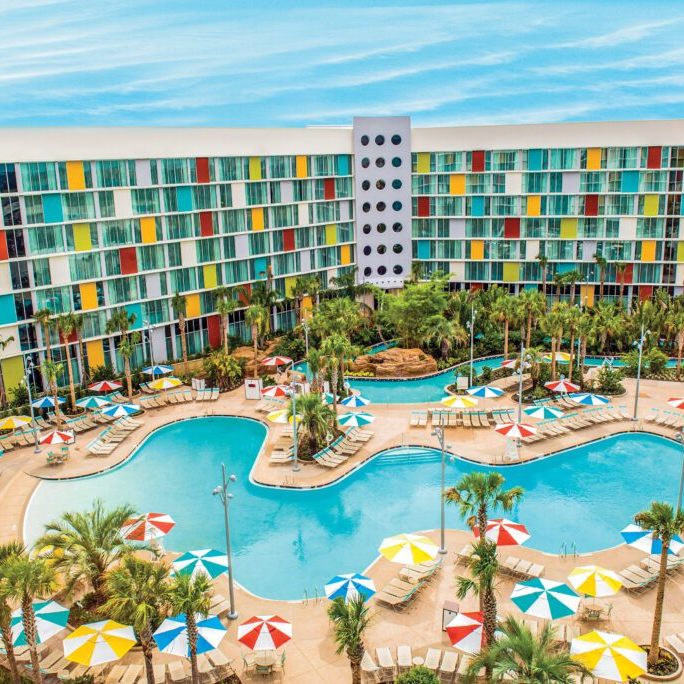 Colorful hotel building surrounding a large, inviting pool area with palm trees and vibrant umbrellas scattered around.