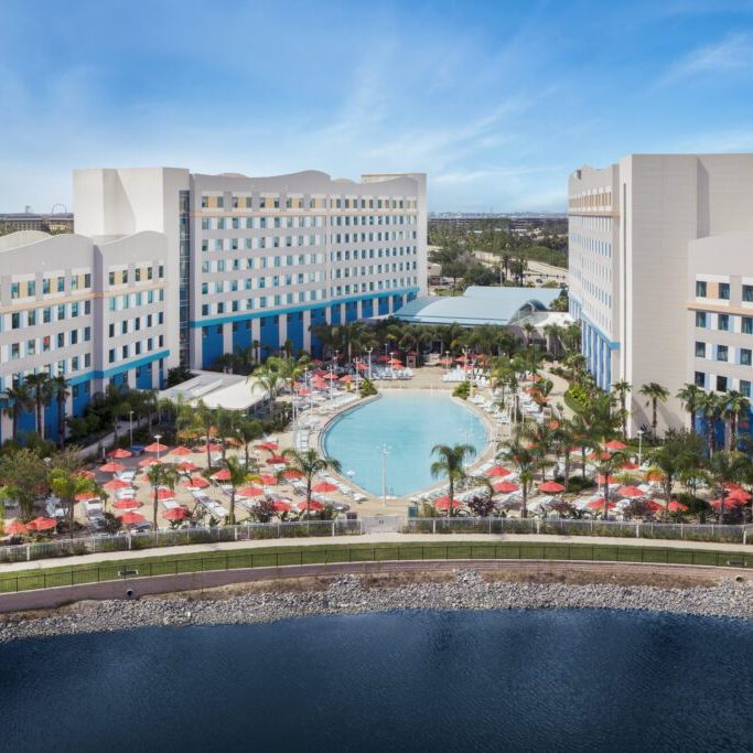 Aerial view of a hotel with a large pool surrounded by palm trees and lounge chairs, next to a calm water body under a blue sky.