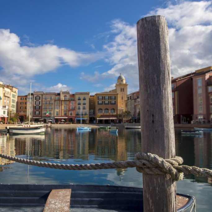 A peaceful waterfront scene featuring colorful buildings, a calm marina, and wooden posts with a rope, under a partly cloudy sky.