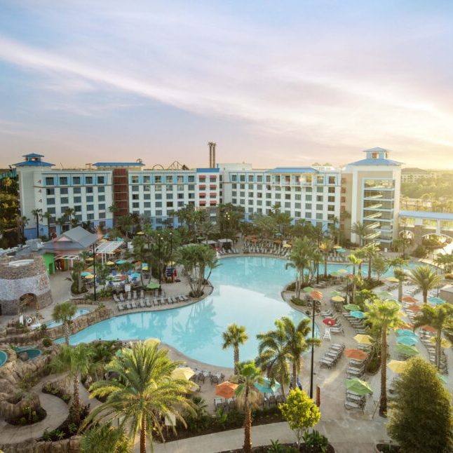 Aerial view of a resort hotel with a large pool and palm trees, set against a sunset sky. The atmosphere is tranquil and inviting.