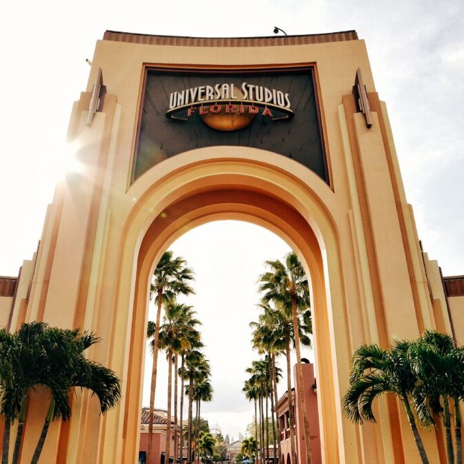 The image features the grand entrance arch of Universal Studios, framed by palm trees and bathed in sunlight.