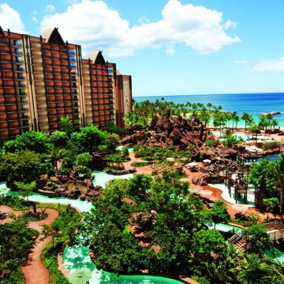 Aerial view of a tropical resort featuring lush gardens, winding pools, and ocean views under a bright blue sky with scattered clouds.